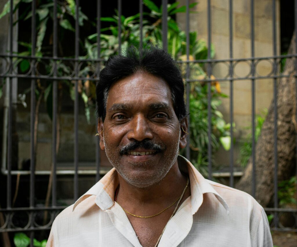 pexels-photo-16447736-16447736 Portrait of a smiling senior Indian man outdoors by a decorative iron fence.